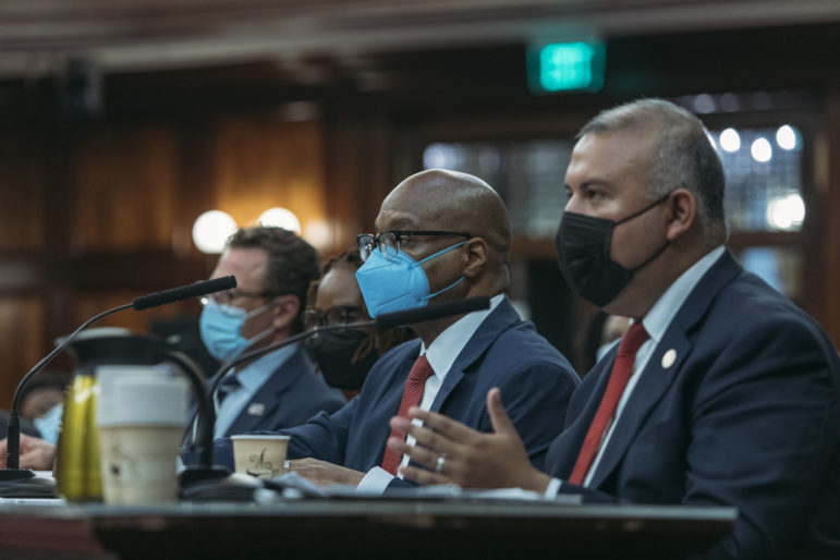 Three men at tables in NYC Council chambers